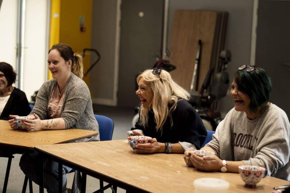 A group of women sat in a row at some tables at The Shack. They are holding cups and laughing.