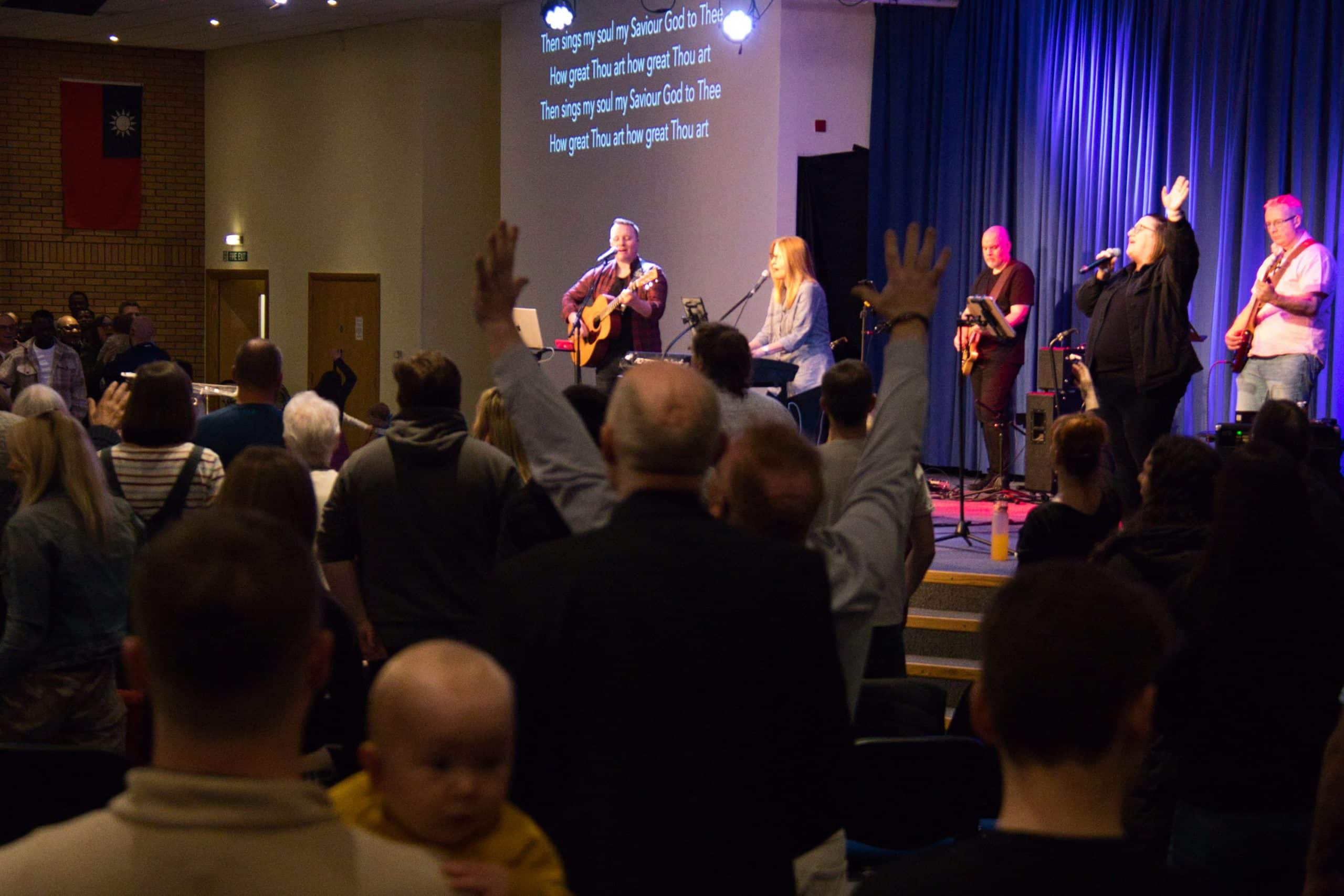 the congregation participating in worship, some with hands in the air, while the worship band plays a song on stage